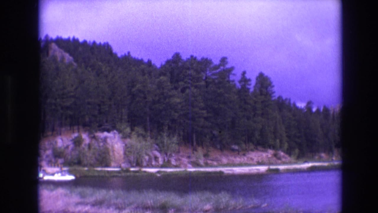 1974 South Dakota 8mm frame of a tree-lined lake with rocky cliffs and a small dock area, including a white boat near the shoreline.