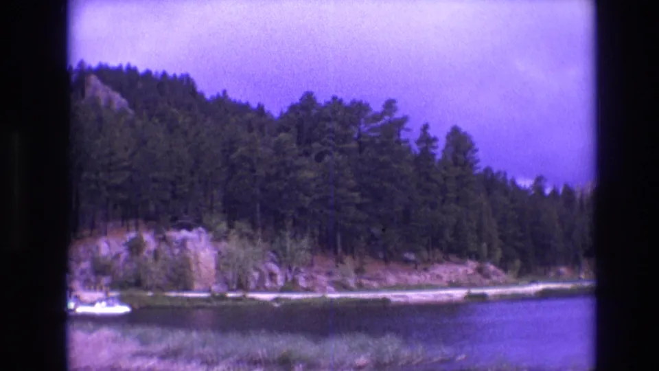 1974 South Dakota 8mm frame of a tree-lined lake with rocky cliffs and a small dock area, including a white boat near the shoreline.