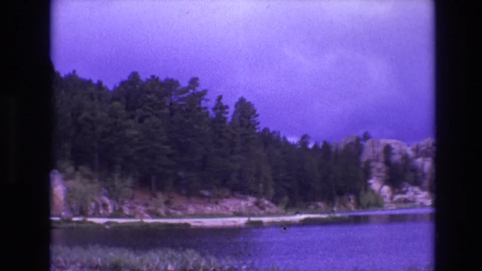 1974 South Dakota 8mm frame showing a wide calm lake bordered by pine forest, pale rocky banks, shoreline reeds, and a purple-tinged cloudy sky.