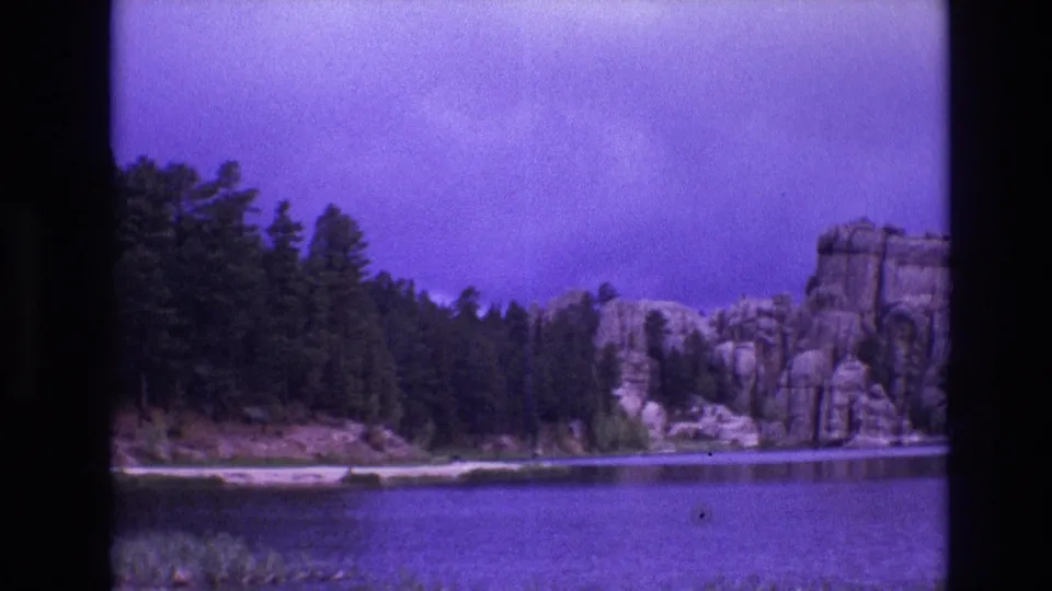 1974 South Dakota 8mm frame with calm lake water in the foreground, pine-covered shore in the middle distance, and large rocky cliffs at the right under a cloudy sky.
