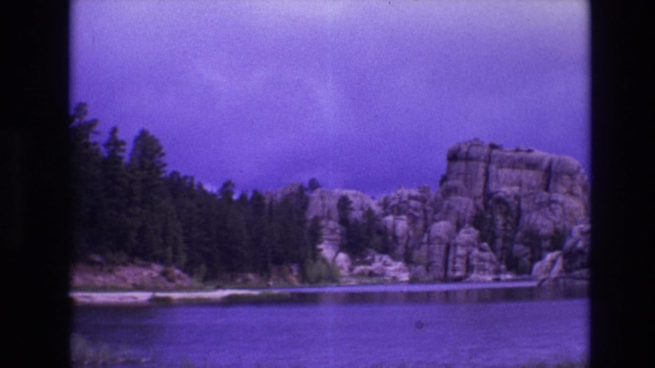1974 South Dakota 8mm frame showing calm lake water, evergreen trees, and large rounded shoreline boulders under a purple-blue sky.