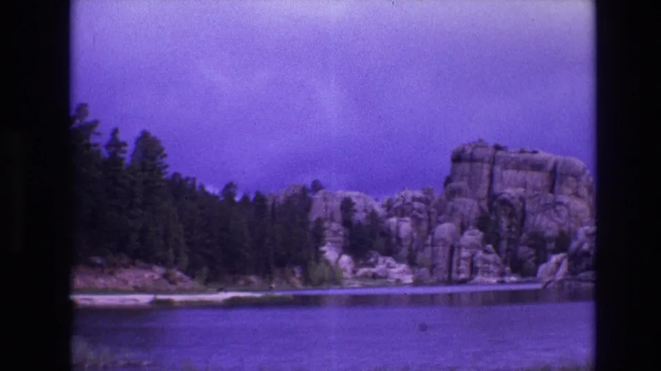 1974 South Dakota 8mm frame showing calm lake water, evergreen trees, and large rounded shoreline boulders under a purple-blue sky.