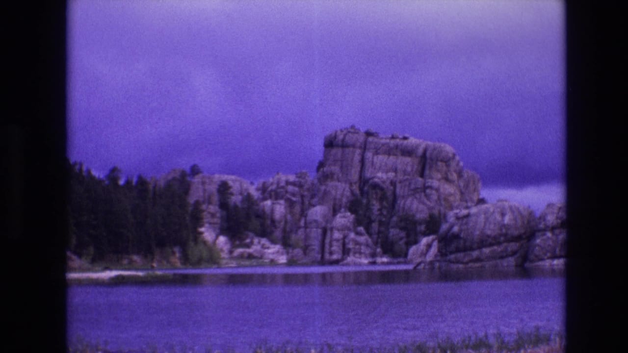 1974 South Dakota 8mm frame with still lake water, a dark tree line, and prominent rounded rock formations along the far shore.
