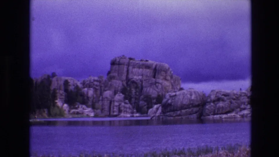 1974 South Dakota 8mm frame looking across calm water toward clustered shoreline boulders, evergreen trees, and a heavy purple-tinted sky.