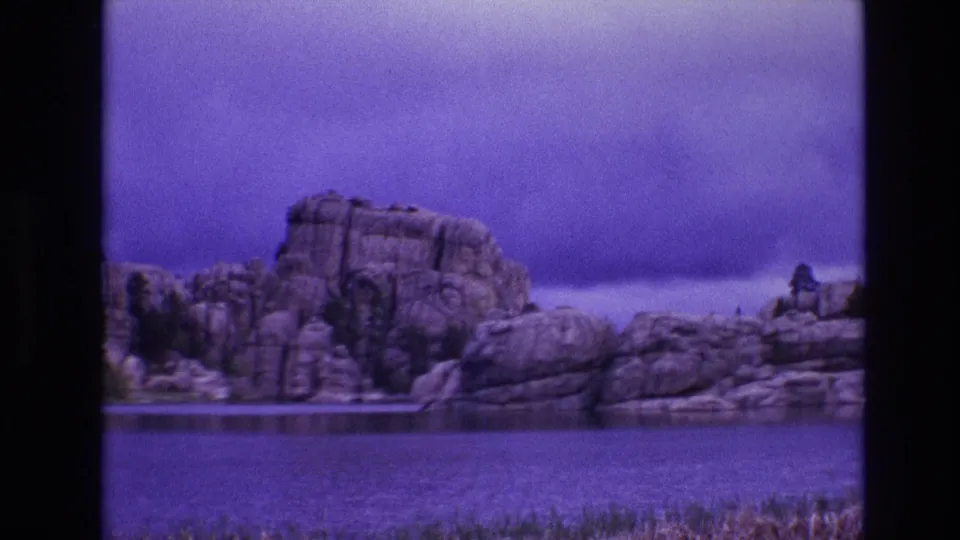 1974 South Dakota 8mm frame showing broad lake water, shoreline rock formations, scattered evergreens, and a purple-tinted overcast sky.