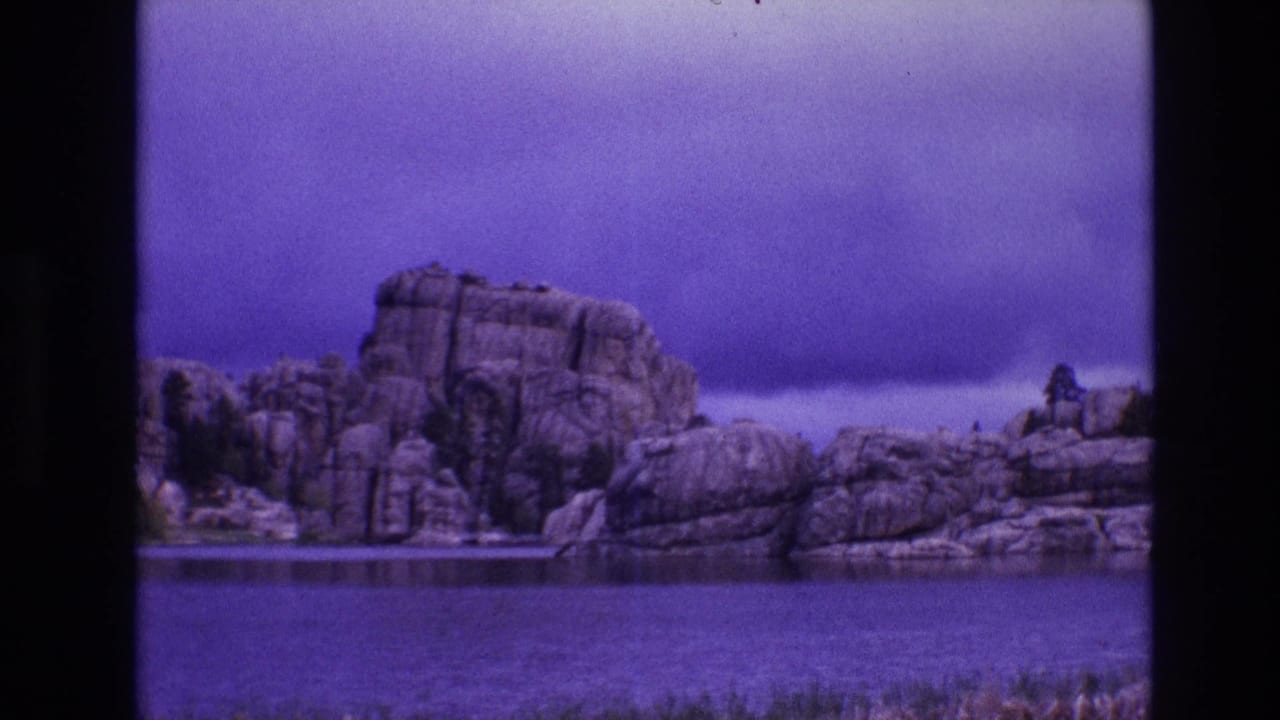 1974 8mm home-movie frame from South Dakota showing a rocky lakeside formation under a purple-blue sky, viewed through dark film borders.