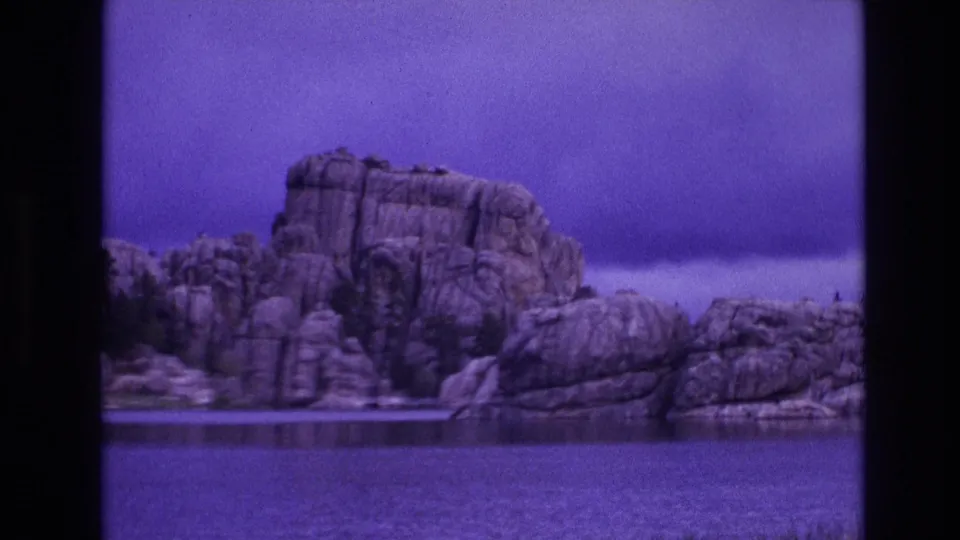 South Dakota 8mm travel footage frame showing a rugged rock formation beside still water with a strong blue-purple color shift.