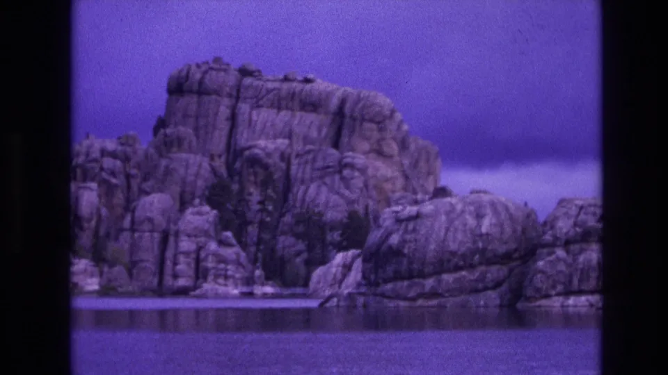 1974 South Dakota home-movie frame with a closer view of vertical rock faces, sparse vegetation, and lake water in the foreground.
