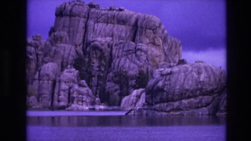 South Dakota 8mm frame showing a large rugged rock formation beside calm lake water, with trees or shrubs visible in the rock crevices.