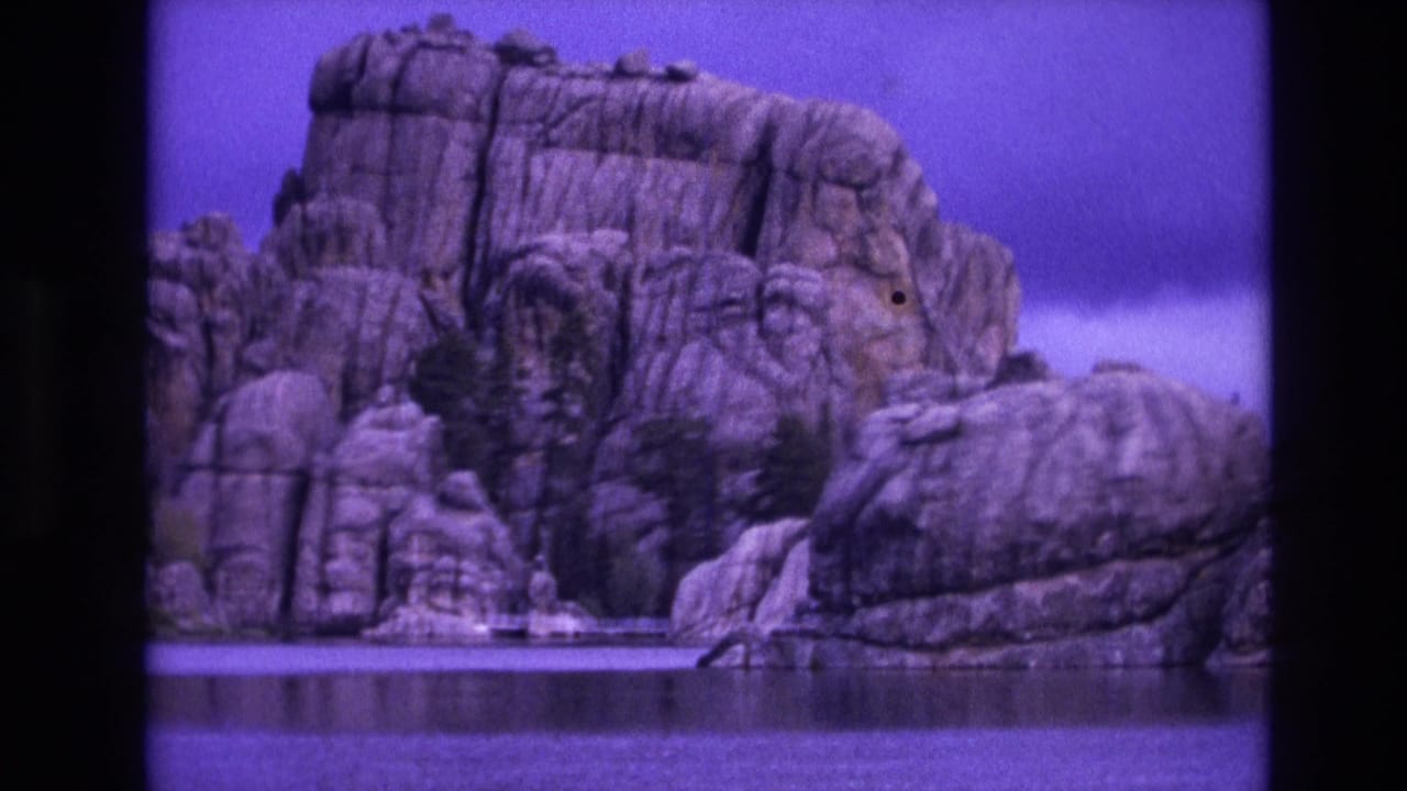 1974 South Dakota 8mm frame showing a rugged rock formation across calm water, with a strong purple color cast and visible film borders.