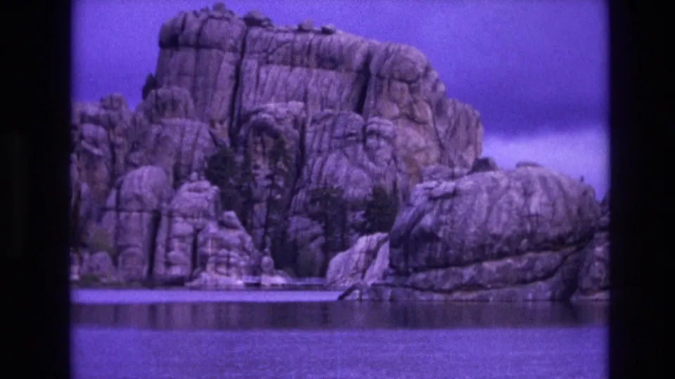 1974 South Dakota 8mm frame with a broad granite-like rock face, foreground water, and analog grain beneath a purple-tinted sky.