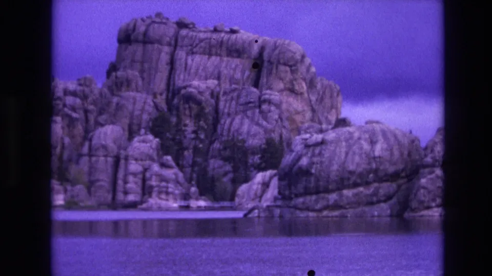 1974 South Dakota home-movie frame showing the same waterside rock formation, with dark frame edges, specks, and a saturated purple cast.