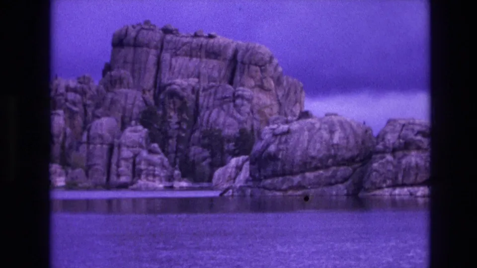 1974 South Dakota 8mm frame showing a rocky shoreline and large rock outcrop across still water, with visible grain and purple color shift.