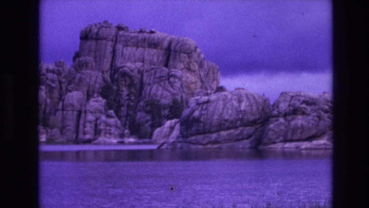 1974 South Dakota 8mm frame showing a rugged lakeside rock formation beyond calm water, with grass at the shore and a strong purple film cast.
