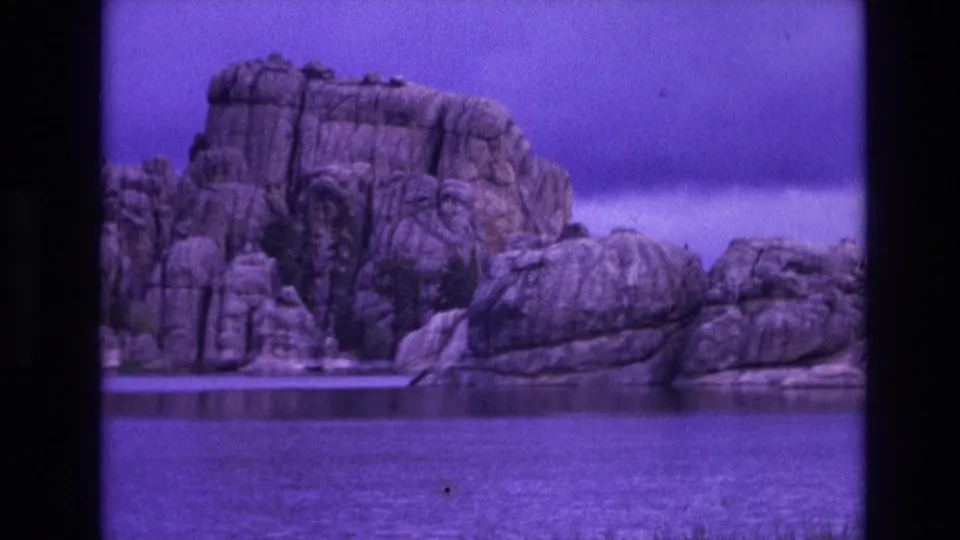 1974 South Dakota 8mm frame showing a rugged lakeside rock formation beyond calm water, with grass at the shore and a strong purple film cast.