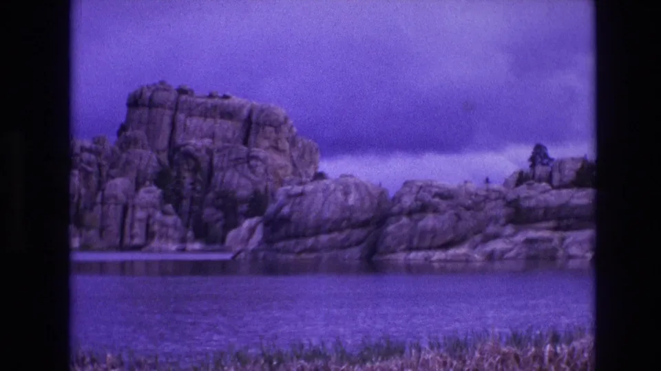 1974 South Dakota 8mm frame showing a rocky shoreline across a lake, with grass along the lower edge and a soft purple cast over the landscape.