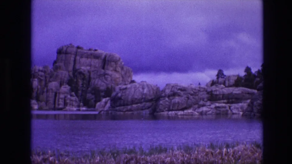 1974 South Dakota frame from 8mm home-movie footage showing a broad view of a rock formation at a lake, with shoreline vegetation and purple-toned sky and water.