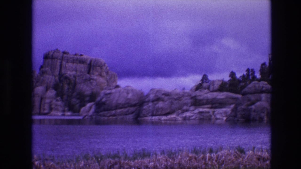 1974 South Dakota 8mm frame showing a purple-toned lakeside view with rounded rocks, pine trees, still water, and cloudy sky.