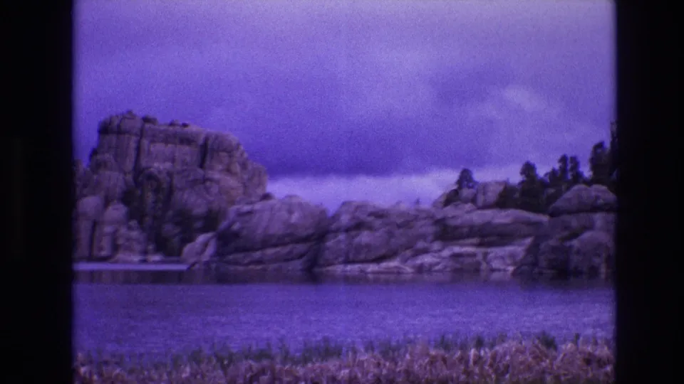 1974 South Dakota 8mm frame showing a purple-toned lakeside view with rounded rocks, pine trees, still water, and cloudy sky.