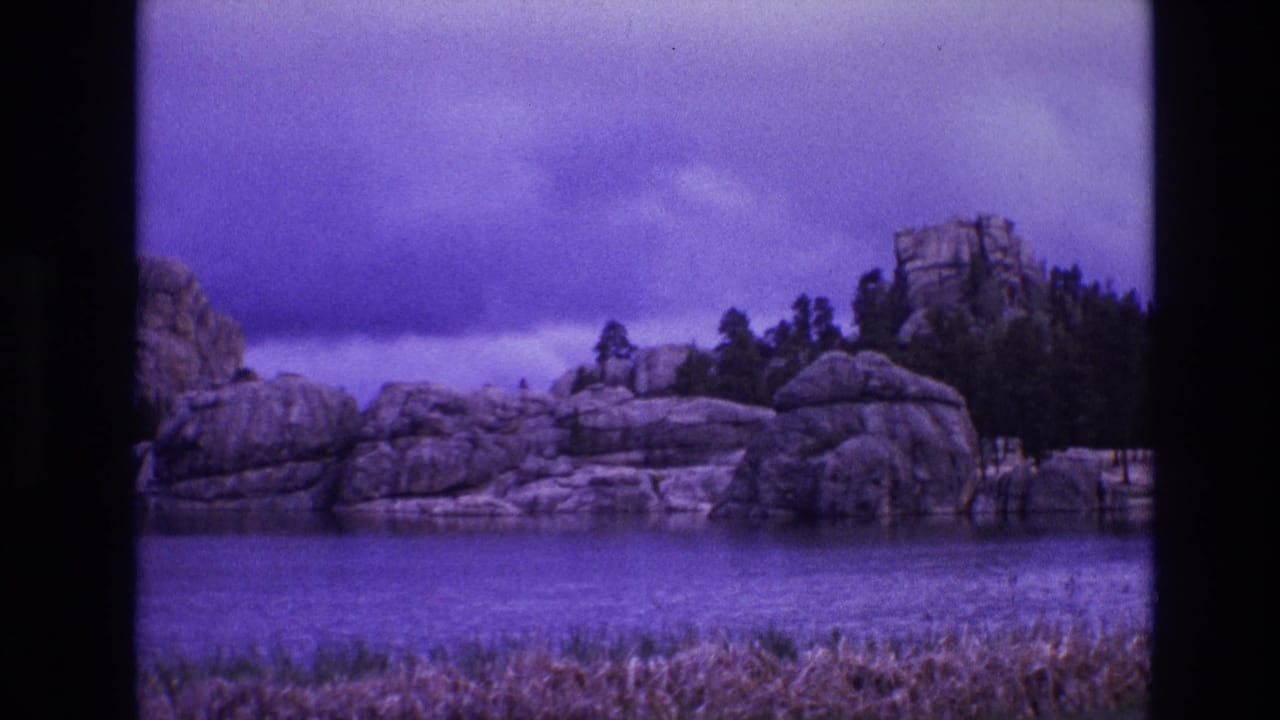 Rocky shoreline across water with pine trees and a purple cloudy sky.