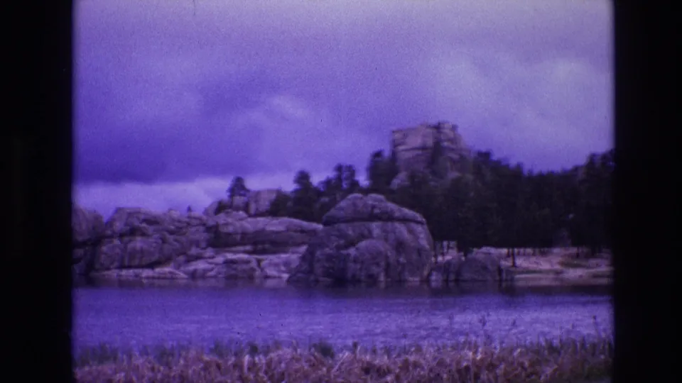 1974 South Dakota 8mm frame with dry grass in the foreground, water, rounded shoreline rocks, pines, and a purple cast across the sky.