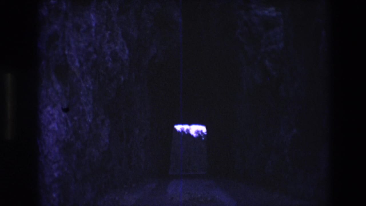 1974 South Dakota 8mm frame showing a dim cave-like stone interior with a small purple-lit opening or fixture set back in shadow.