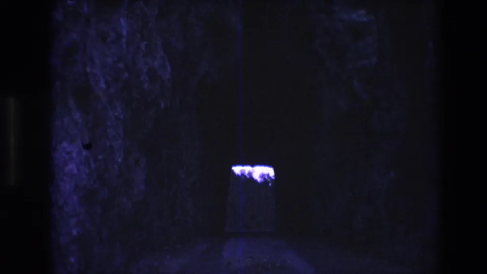 1974 South Dakota 8mm frame showing a dim cave-like stone interior with a small purple-lit opening or fixture set back in shadow.