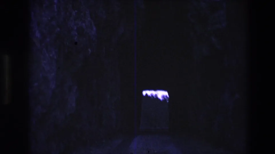 1974 South Dakota 8mm frame of a shadowed cave-like wall with a small bright purple-lit opening or fixture to the right.