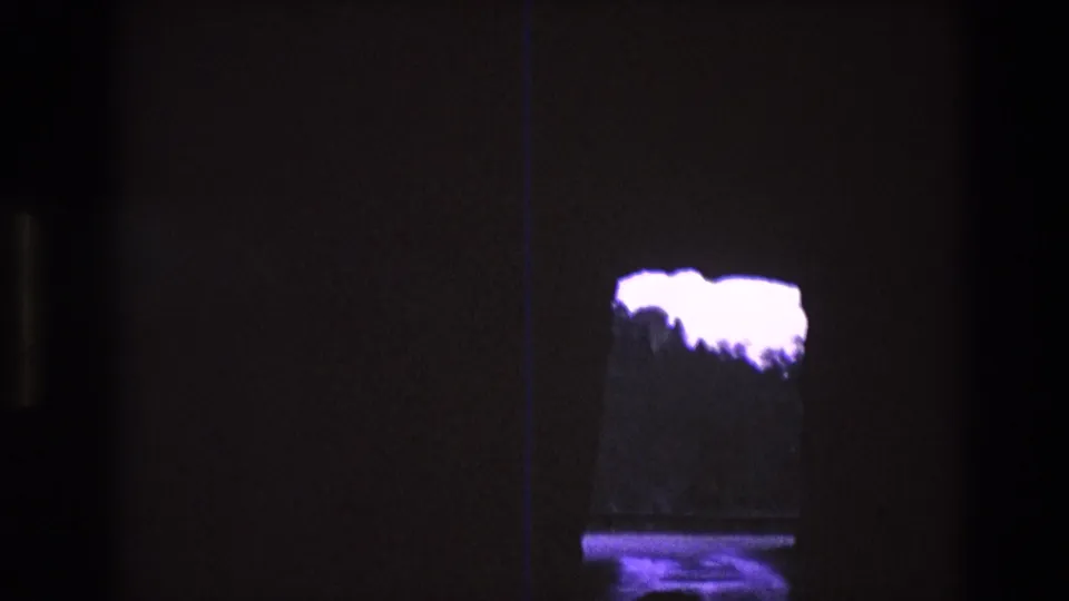 1974 South Dakota 8mm frame looking from a dark cave-like opening toward overexposed purple sky, a distant treeline, and a reflective surface below.