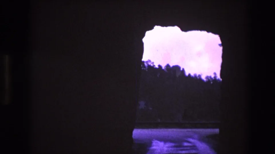 1974 South Dakota 8mm frame showing a larger cave-like opening with pale purple overexposed sky, a dark tree ridge, and possible water reflection below.