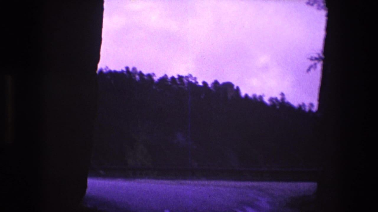 A dark-edged 8mm frame shows a paved road curving past wooded hills in South Dakota countryside, with no people visible.