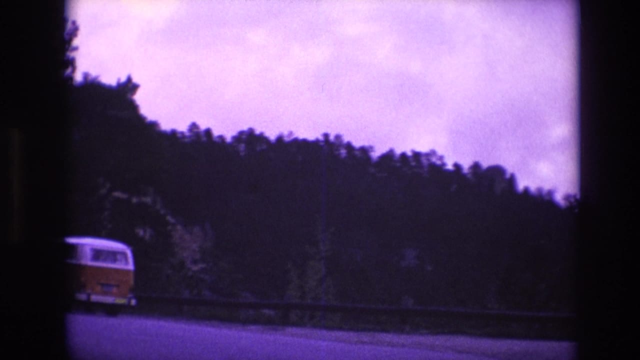 A yellow-and-white van travels along a winding highway bordered by a guardrail and forested hills in the 1974 South Dakota home-movie footage.