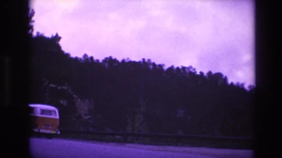 A yellow-and-white van travels along a winding highway bordered by a guardrail and forested hills in the 1974 South Dakota home-movie footage.