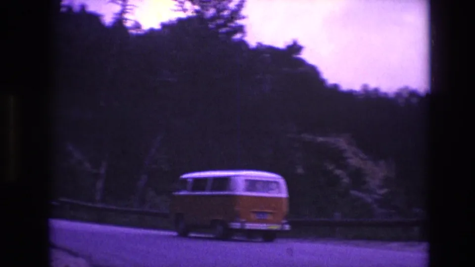 The yellow-and-white van rounds a highway bend beneath dark trees and wooded hills, with soft 8mm focus and strong purple tint.