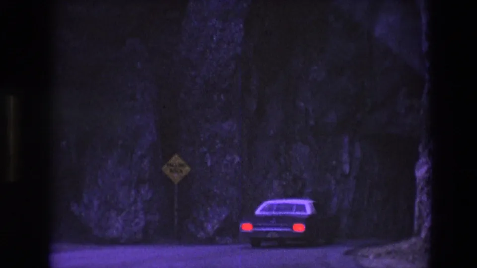 A car with red taillights appears near a roadside warning sign and dark rock-lined opening or tunnel entrance along the South Dakota highway.