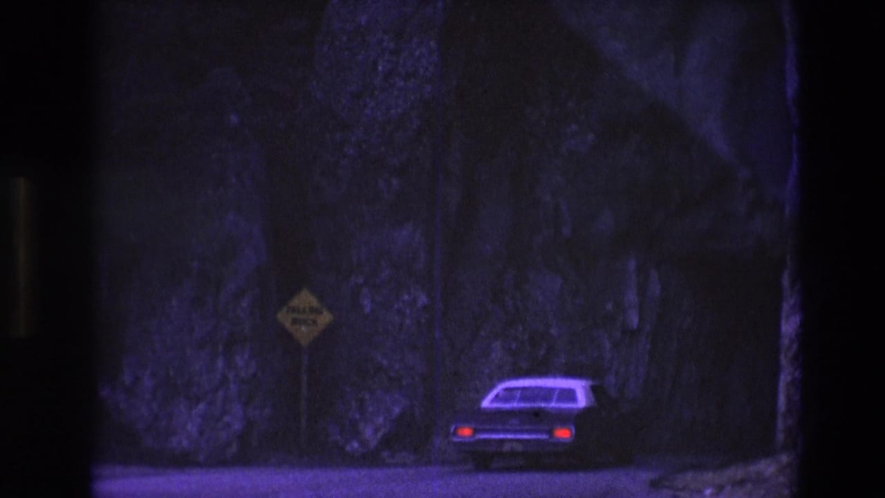 An older car drives away through a shadowed South Dakota mountain pass in 1974 8mm footage, with a yellow falling-rock sign near the rocky roadside.