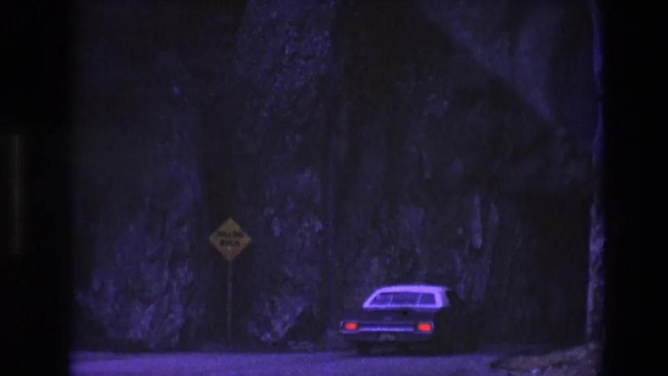 An older car drives away through a shadowed South Dakota mountain pass in 1974 8mm footage, with a yellow falling-rock sign near the rocky roadside.