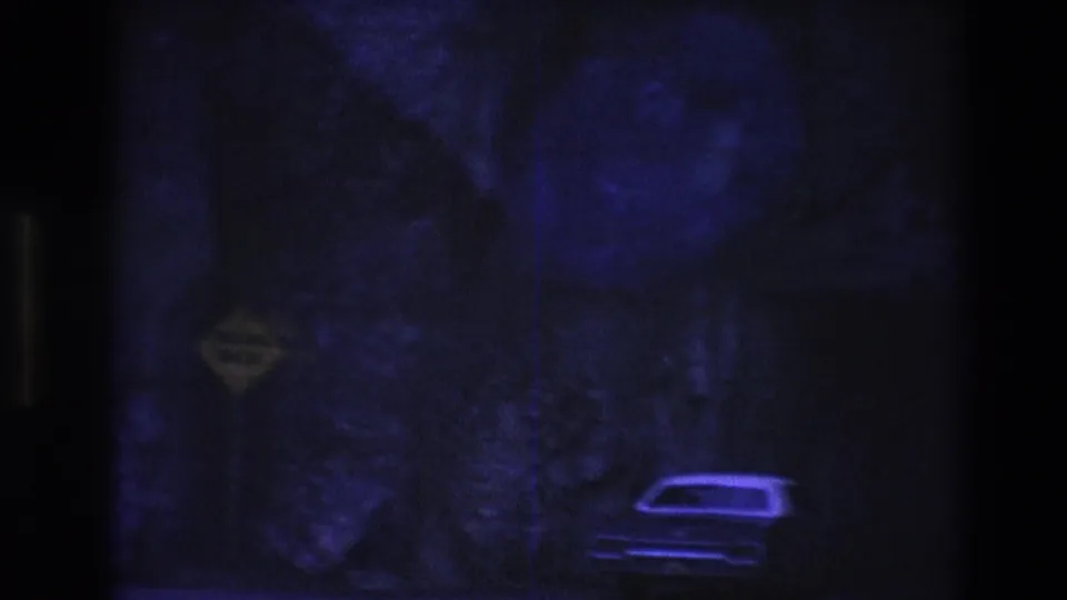 In a heavily shadowed 1974 8mm frame, an older car sits low in the composition beneath a rocky wall and a yellow falling-rock warning sign.
