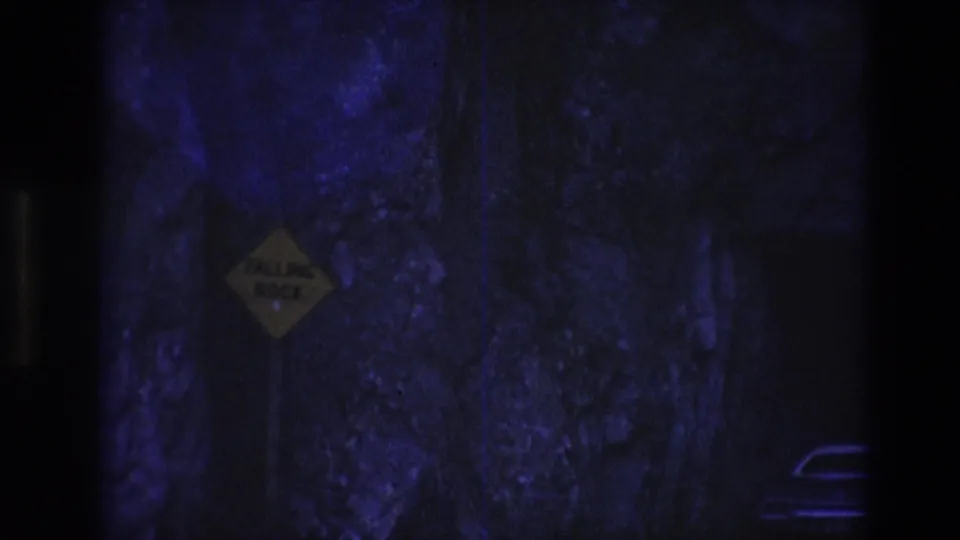 1974 South Dakota 8mm frame showing a dim rock-walled tunnel or passageway with blue-purple lighting and a yellow diamond warning sign on the left.