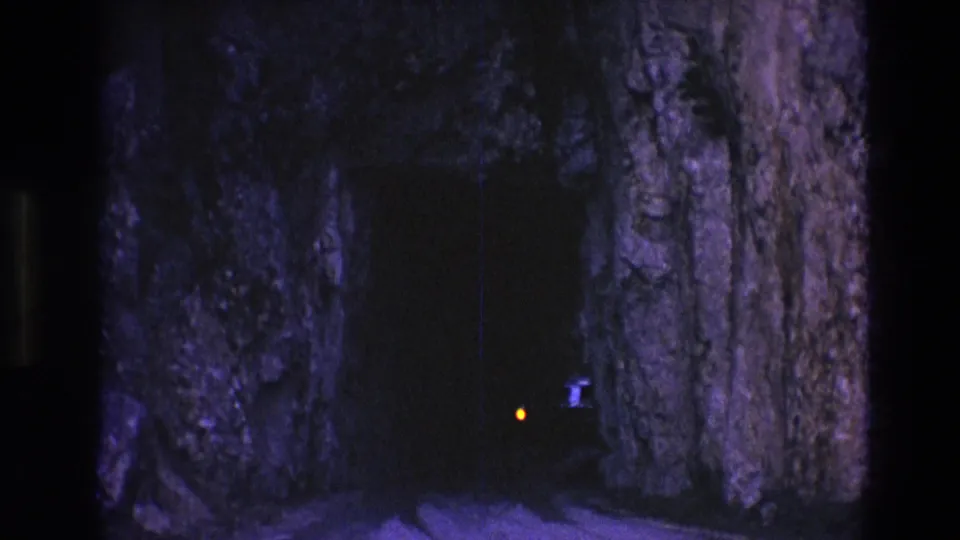 Final 1974 South Dakota frame with a small vehicle light beside a shadowed tunnel opening and rough illuminated rock walls.