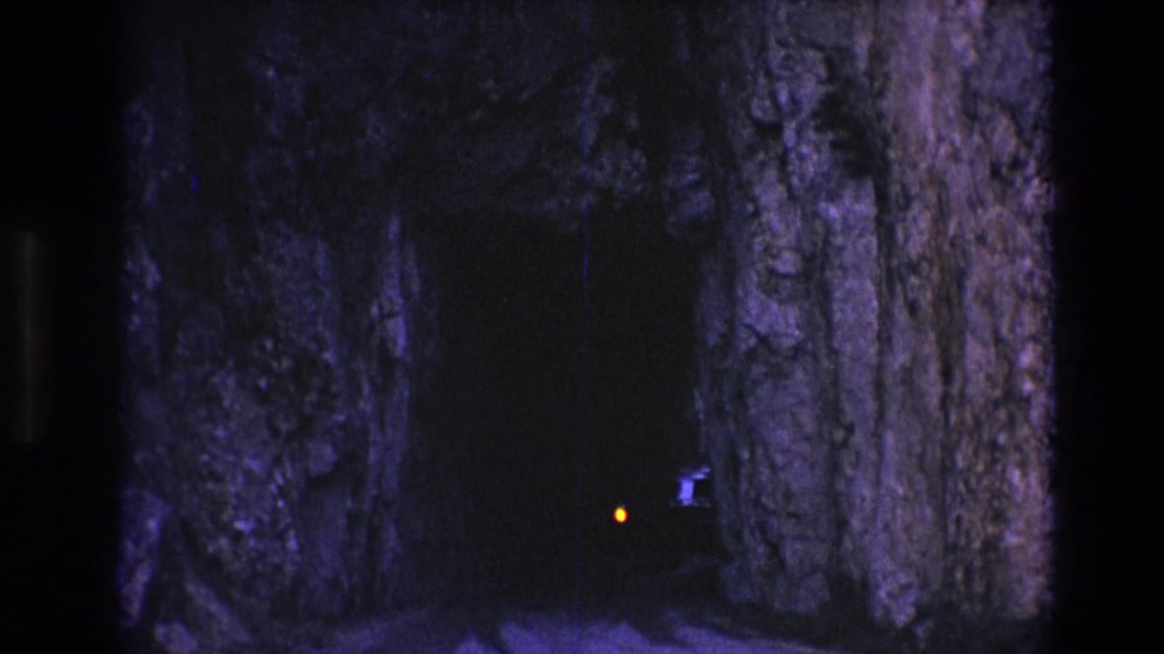 1974 South Dakota 8mm frame showing a dark mountain tunnel or rock passage with purple-blue rock walls and a small orange light approaching from inside.