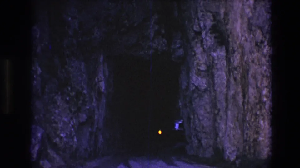 1974 South Dakota 8mm frame showing a dark mountain tunnel or rock passage with purple-blue rock walls and a small orange light approaching from inside.