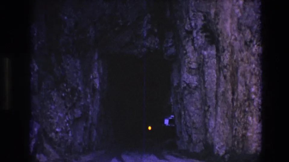 1974 South Dakota 8mm frame with a small orange train light and faint dark form emerging beside purple-toned rock inside a tunnel.