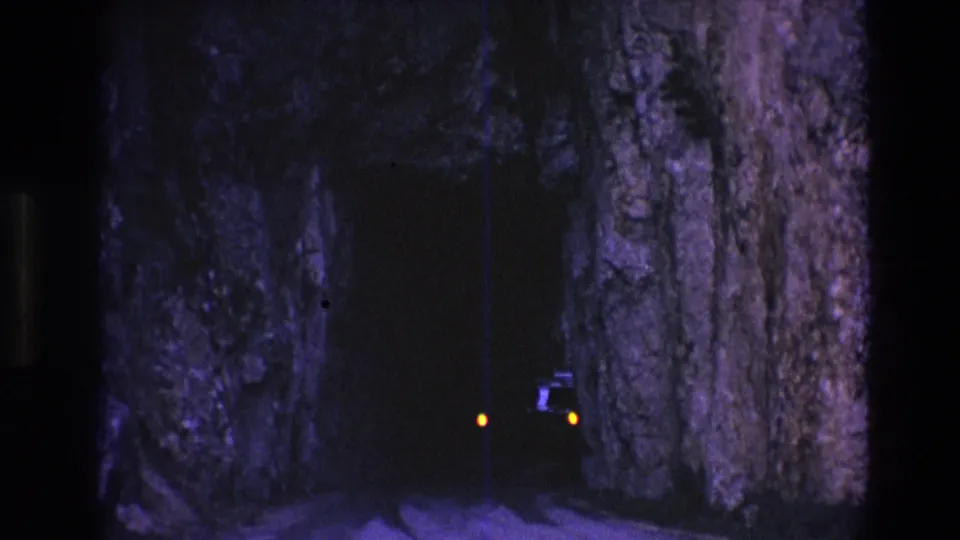 1974 South Dakota 8mm frame with orange train lights glowing in a shadowed rock tunnel, framed by purple-blue stone and visible film wear.