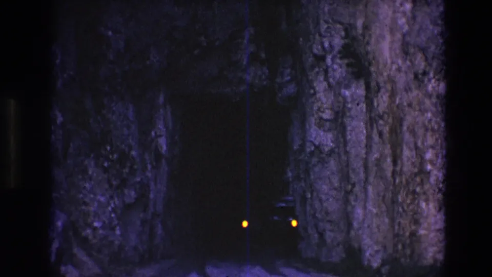 1974 home-movie frame showing a car partly emerging from a narrow South Dakota rock tunnel, with one amber light and the vehicle edge visible in darkness.