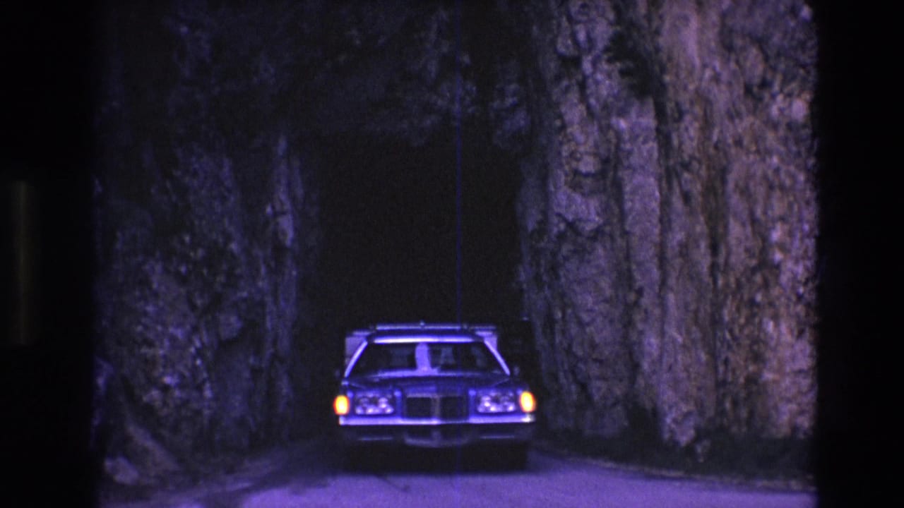 A 1970s station wagon appears head-on in a dark, narrow South Dakota rock tunnel, with headlights glowing against rough stone walls.