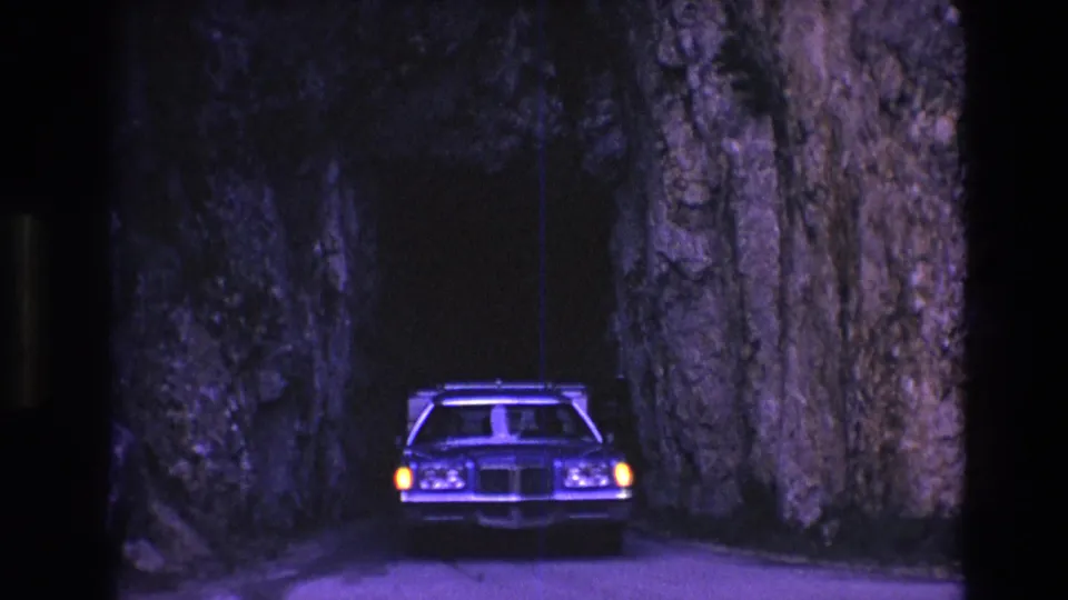 A 1970s station wagon appears head-on in a dark, narrow South Dakota rock tunnel, with headlights glowing against rough stone walls.