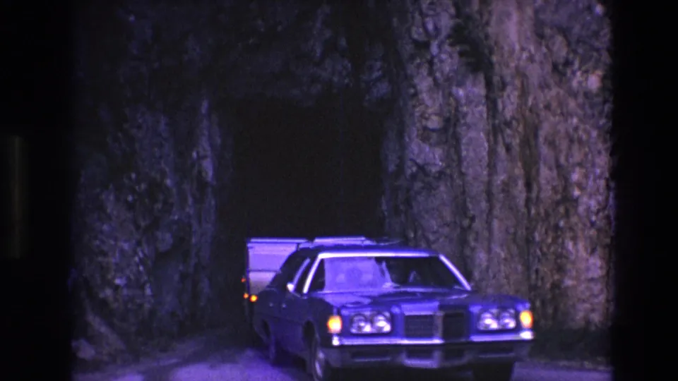 The station wagon moves toward the tunnel exit in South Dakota, with a light-colored camper trailer visible behind it against the rock wall.