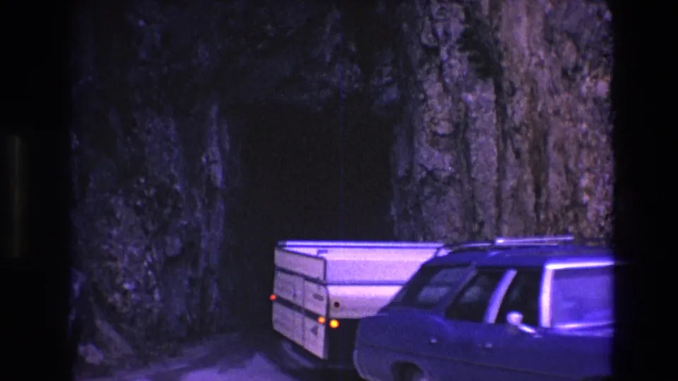 The light-colored camper trailer and rear side of the station wagon pass close to the camera beside the rocky South Dakota tunnel wall.