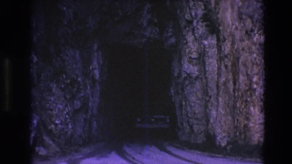 1974 South Dakota home-movie frame showing a light-colored car near a narrow roadway cut through tall rock walls.